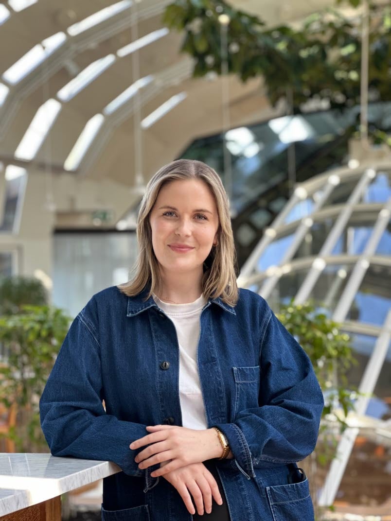 Kvinna i mörkblont hår, jeansjacka och vit t-shirt. Kvinnan står lutad mot ett marmorbord